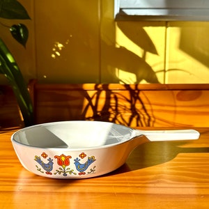 May include: White ceramic frying pan with a blue and orange floral design featuring birds. The pan has a black handle.