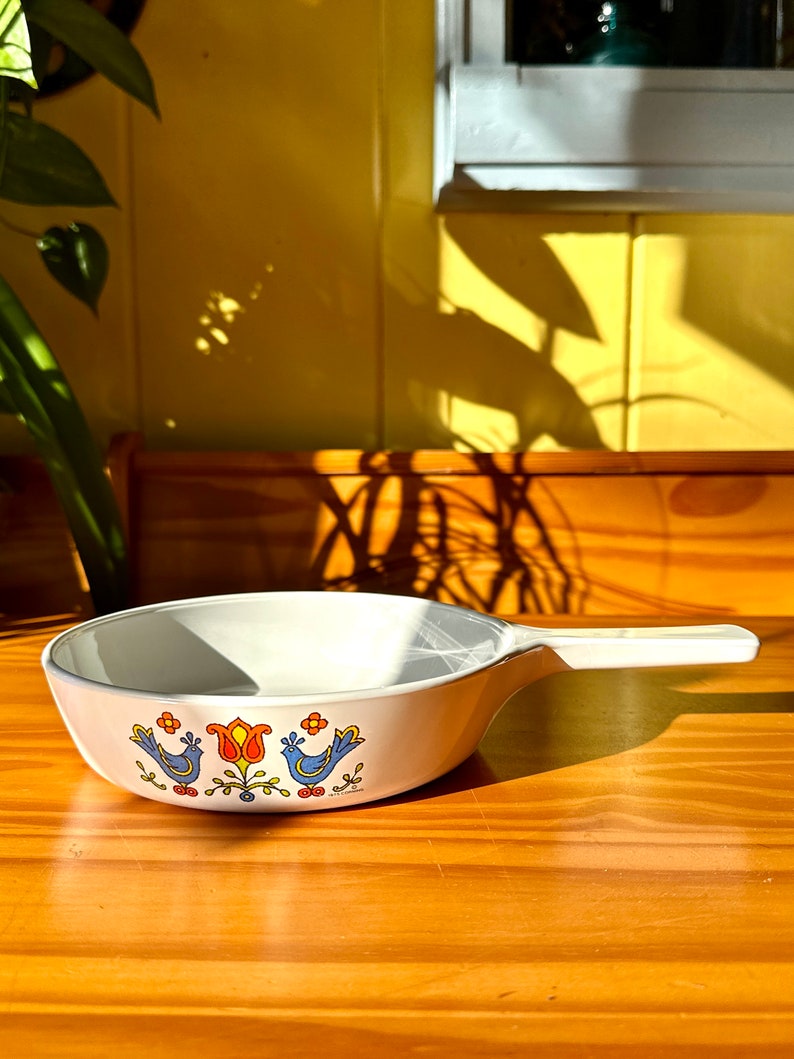 May include: White ceramic frying pan with a blue and orange floral design featuring birds. The pan has a black handle.