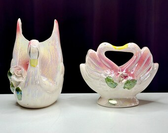 May include: Two white ceramic swan figurines with pink and green accents. One swan is standing upright with its neck curved, while the other is in a heart shape with its necks intertwined.