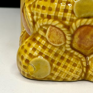 May include: Close-up of a ceramic object with a yellow and brown checkered pattern. The object has a rounded shape with several brown, oval-shaped accents. The base of the object is visible, showing a small chip in the glaze.