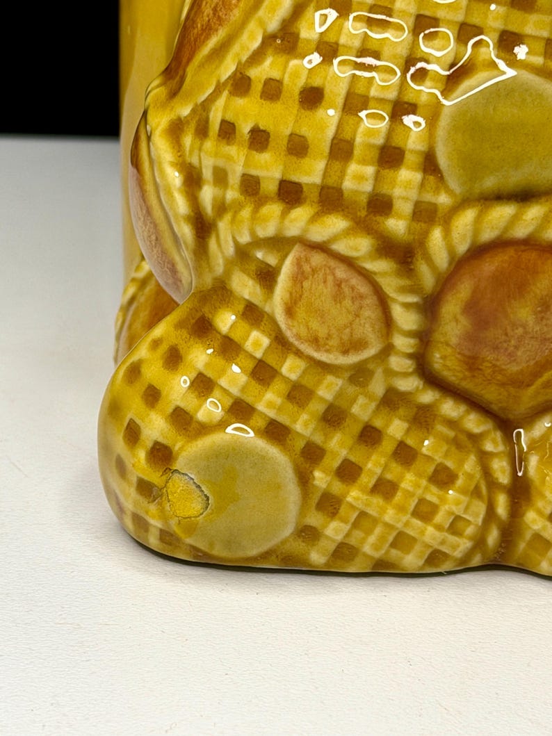 May include: Close-up of a ceramic object with a yellow and brown checkered pattern. The object has a rounded shape with several brown, oval-shaped accents. The base of the object is visible, showing a small chip in the glaze.