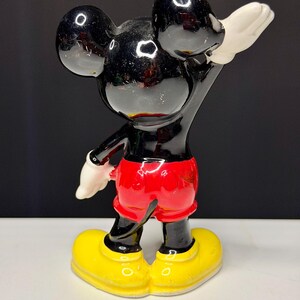 K&ouml;nnte beinhalten: Eine Vintage-Mickey-Mouse-Figur. Die schwarze Figur hat gro&szlig;e runde Ohren, rote Shorts, gelbe Schuhe und wei&szlig;e Handschuhe. Die Figur posiert mit einem erhobenen Arm. Das gl&auml;nzende Finish reflektiert das Licht.
