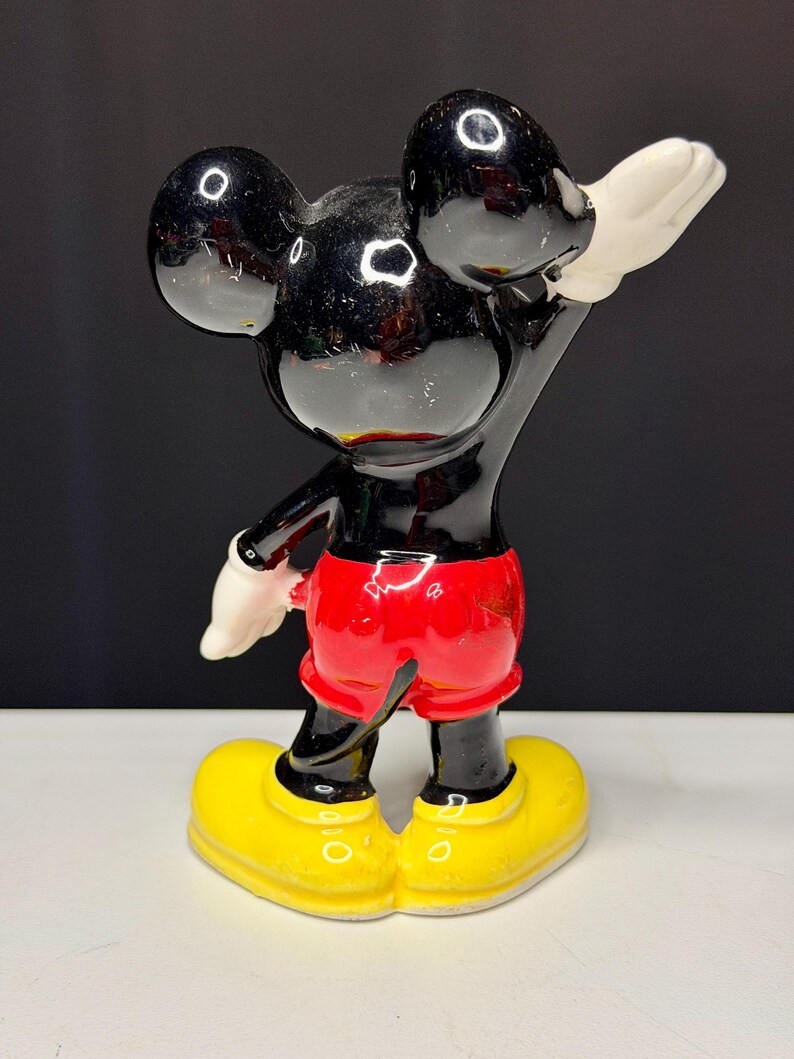 K&ouml;nnte beinhalten: Eine Vintage-Mickey-Mouse-Figur. Die schwarze Figur hat gro&szlig;e runde Ohren, rote Shorts, gelbe Schuhe und wei&szlig;e Handschuhe. Die Figur posiert mit einem erhobenen Arm. Das gl&auml;nzende Finish reflektiert das Licht.
