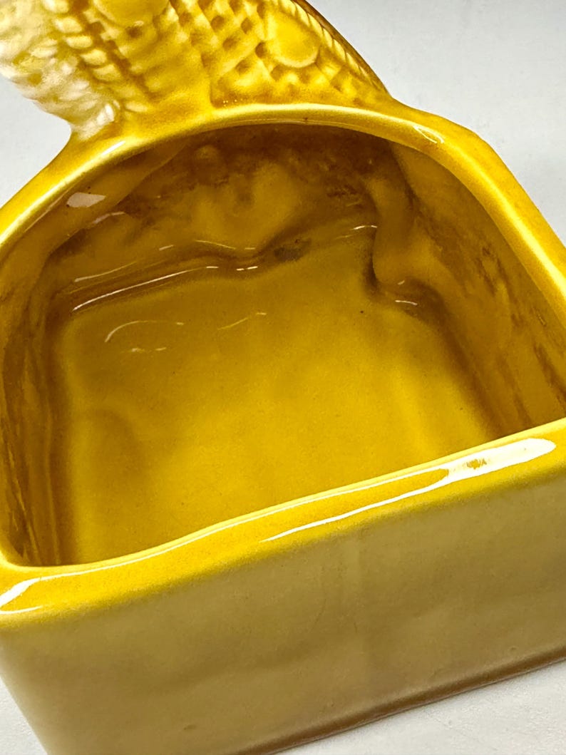 May include: A close-up of a yellow ceramic dish with a bird-shaped handle. The dish has a rectangular shape with rounded corners and a glossy finish. The interior is a darker shade of yellow.
