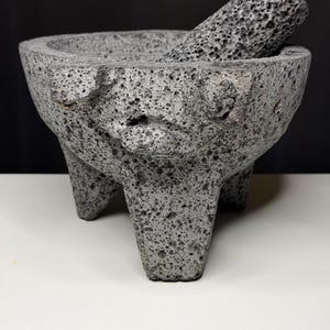 May include: A gray, three-legged molcajete and tejolote set. The mortar has a wide bowl and the pestle is cylindrical. The porous volcanic rock has a rough texture. The set is designed for grinding spices and making sauces.