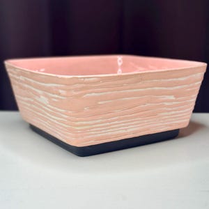 May include: A square, pink ceramic planter with a textured, wood-like design. The planter sits on a black base. The pink color is a soft, muted tone, and the white lines add contrast. The planter is suitable for indoor or outdoor use.
