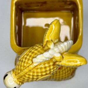 May include: A yellow ceramic butter dish with a corn-shaped handle. The dish is rectangular with rounded corners. The corn handle has a detailed, textured design with white accents and black eyes.