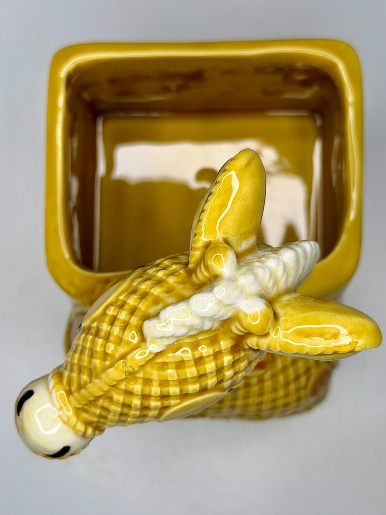 May include: A yellow ceramic butter dish with a corn-shaped handle. The dish is rectangular with rounded corners. The corn handle has a detailed, textured design with white accents and black eyes.