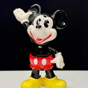 K&ouml;nnte beinhalten: Eine Keramikfigur von Mickey Mouse, einer klassischen Disney-Figur. Mickey wird mit schwarzem K&ouml;rper, wei&szlig;en Handschuhen und gelben Schuhen dargestellt. Er tr&auml;gt rote Shorts mit wei&szlig;en Kn&ouml;pfen und hat einen fr&ouml;hlichen Gesichtsausdruck.