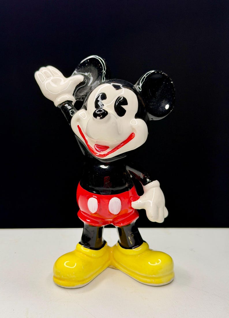 K&ouml;nnte beinhalten: Eine Keramikfigur von Mickey Mouse, einer klassischen Disney-Figur. Mickey wird mit schwarzem K&ouml;rper, wei&szlig;en Handschuhen und gelben Schuhen dargestellt. Er tr&auml;gt rote Shorts mit wei&szlig;en Kn&ouml;pfen und hat einen fr&ouml;hlichen Gesichtsausdruck.