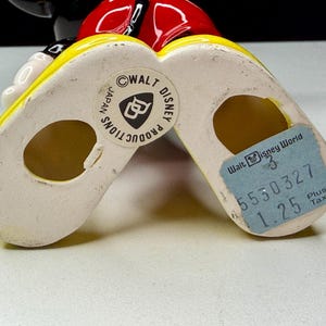 K&ouml;nnte beinhalten: Vintage Mickey Mouse Figur, von den F&uuml;&szlig;en aus gesehen, mit gelben Schuhen und wei&szlig;er Basis. Die Figur hat ein rotes Oberteil und einen schwarzen Kopf. Ein blaues Etikett lautet "Walt Disney World". Markiert mit "Walt Disney Productions Japan".