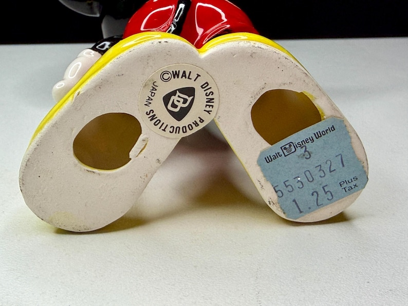 K&ouml;nnte beinhalten: Vintage Mickey Mouse Figur, von den F&uuml;&szlig;en aus gesehen, mit gelben Schuhen und wei&szlig;er Basis. Die Figur hat ein rotes Oberteil und einen schwarzen Kopf. Ein blaues Etikett lautet "Walt Disney World". Markiert mit "Walt Disney Productions Japan".