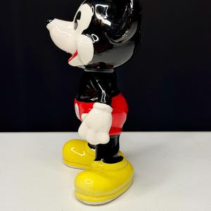 K&ouml;nnte beinhalten: Eine Keramikfigur von Micky Maus. Die Figur ist im Profil zu sehen und zeigt ihren schwarzen Kopf und ihre Ohren, ihr wei&szlig;es Gesicht und ihre roten Shorts. Sie hat gelbe Schuhe und wei&szlig;e Handschuhe. Die Figur ist gl&auml;nzend und steht auf einer wei&szlig;en Oberfl&auml;che vor einem schwarzen Hintergrund.