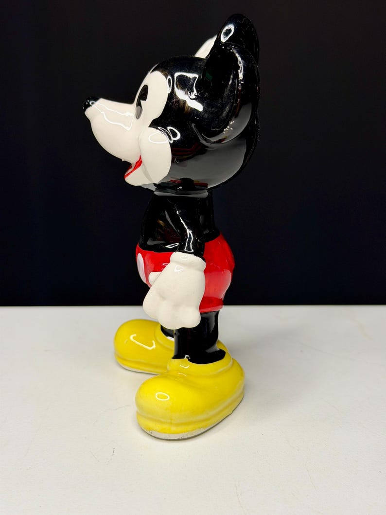 K&ouml;nnte beinhalten: Eine Keramikfigur von Micky Maus. Die Figur ist im Profil zu sehen und zeigt ihren schwarzen Kopf und ihre Ohren, ihr wei&szlig;es Gesicht und ihre roten Shorts. Sie hat gelbe Schuhe und wei&szlig;e Handschuhe. Die Figur ist gl&auml;nzend und steht auf einer wei&szlig;en Oberfl&auml;che vor einem schwarzen Hintergrund.