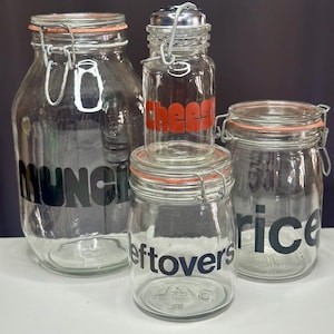 May include: Four clear glass jars with metal closures and orange rubber seals. The jars are labeled with the words "MUNCH", "cheese", "leftovers", and "rice" in various fonts. The jars are arranged on a white surface against a black background.