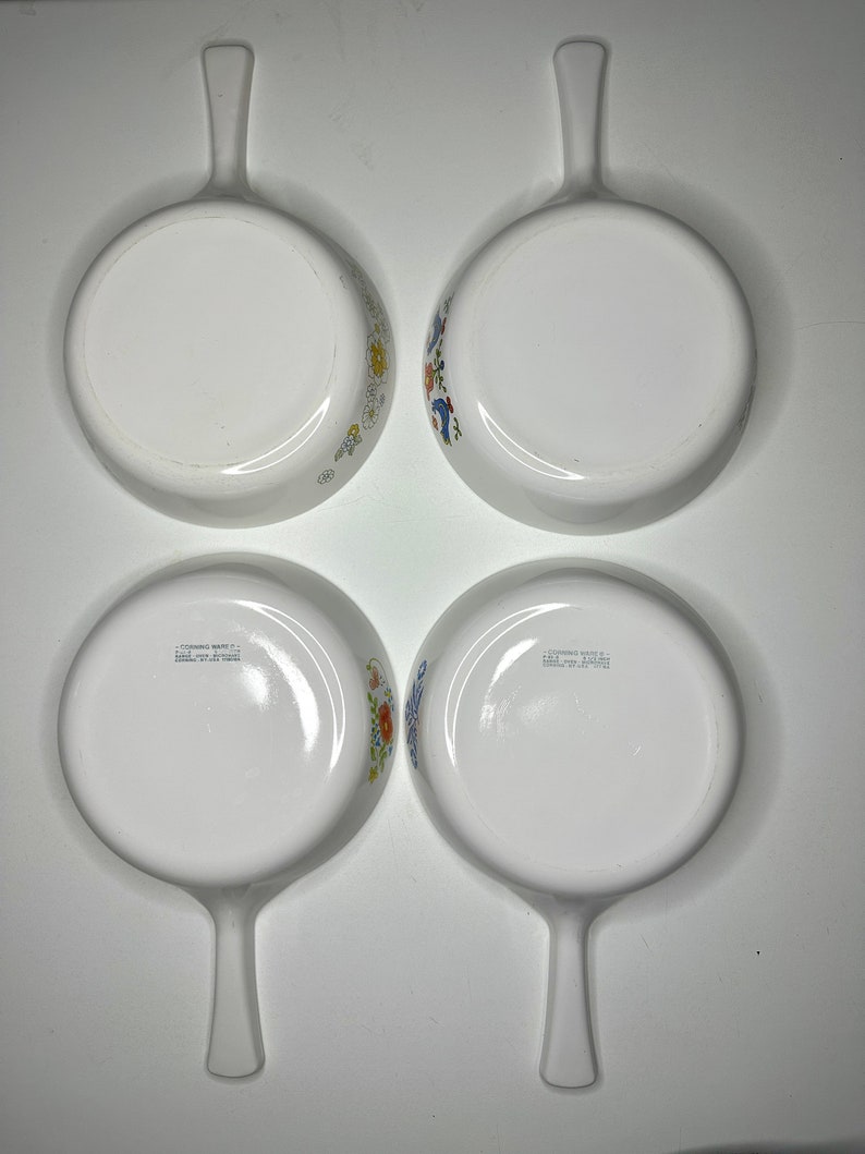 May include: Set of four white ceramic baking dishes with blue and yellow floral designs. Each dish has a single handle and the words "Corning Ware" and "Made in USA" are printed on the bottom.