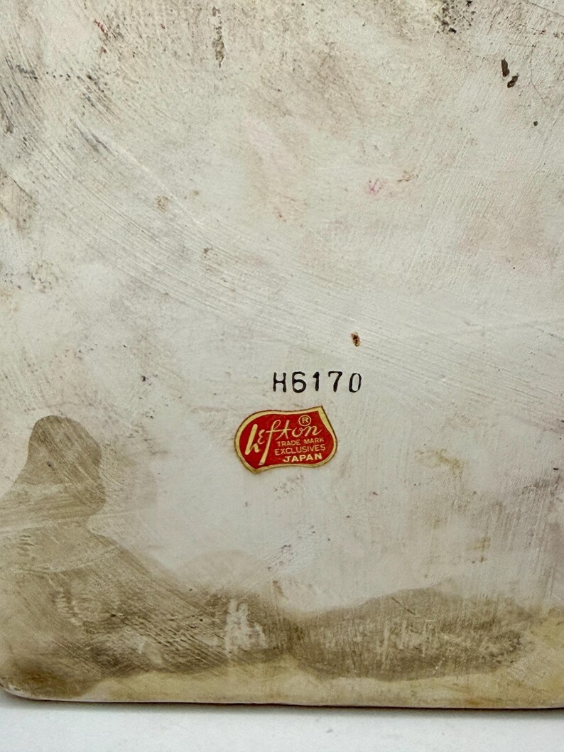 May include: A close-up of a vintage ceramic piece with a textured, off-white surface. The number "H6170" is printed in black. A red and gold "Lefton" sticker is affixed, indicating "Trade Mark Exclusives Japan".