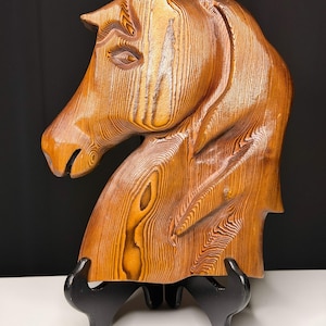 May include: A detailed wooden sculpture of a horse's head, showcasing the grain of the wood. The sculpture is a warm brown color and is mounted on a black stand. The horse's head is in profile, with intricate carved details.