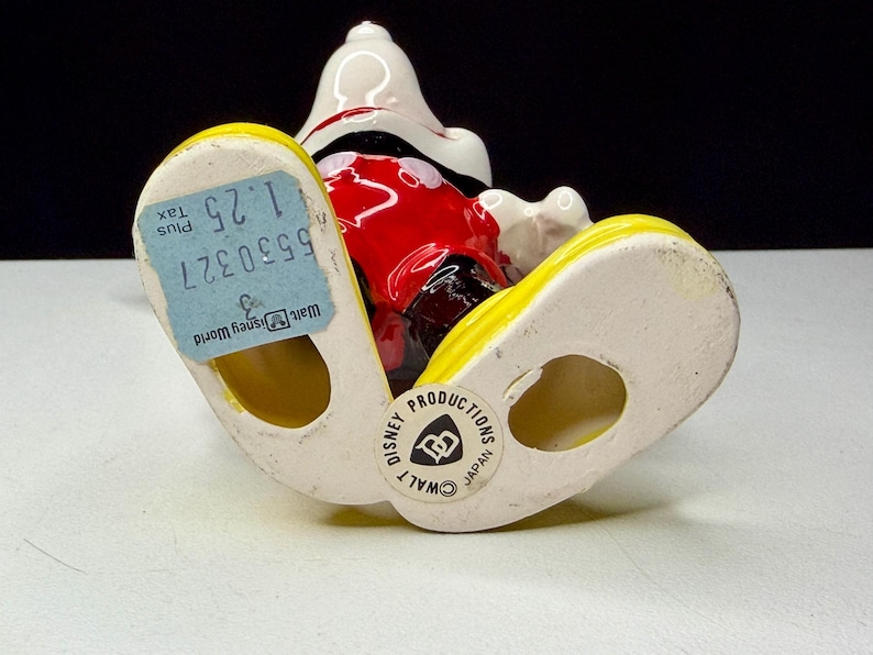K&ouml;nnte beinhalten: Eine Keramikfigur von Mickey Mouse mit wei&szlig;em Kopf, roter Hose und gelben Schuhen. Die Figur hat ein blaues Preisschild an einem Fu&szlig; und einen "Walt Disney World"-Aufkleber am anderen. Die Figur ist mit "Disney Productions Japan" gekennzeichnet.