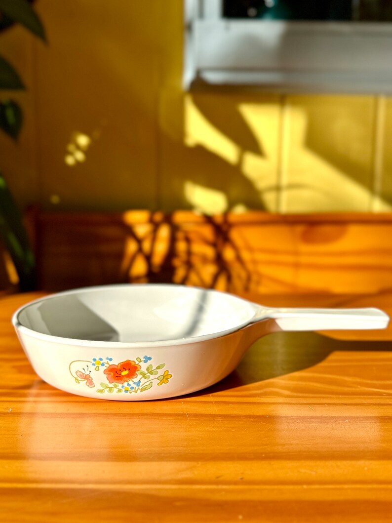 May include: White ceramic saucepan with a floral design featuring orange, red, blue, and green flowers. The saucepan has a white handle.