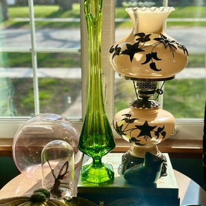 May include: A still life featuring a green glass vase, a white lamp with a floral design, a pink glass orb, a glass dome with a butterfly inside, and two gold butterfly-shaped decorative items.