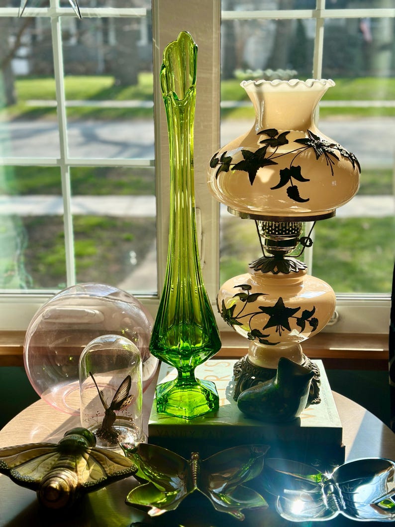 May include: A still life featuring a green glass vase, a white lamp with a floral design, a pink glass orb, a glass dome with a butterfly inside, and two gold butterfly-shaped decorative items.