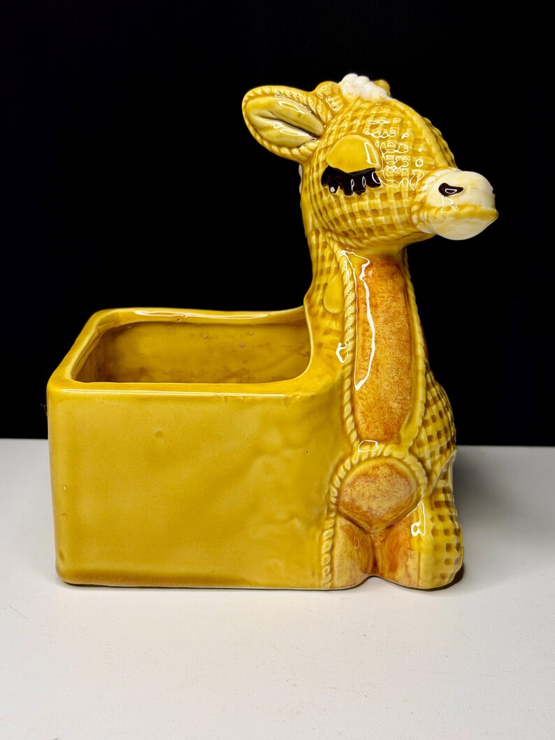 May include: A yellow ceramic giraffe planter with a rectangular container. The giraffe has a textured pattern, black eyes, and a white muzzle. The planter is a decorative item for plants or small items.