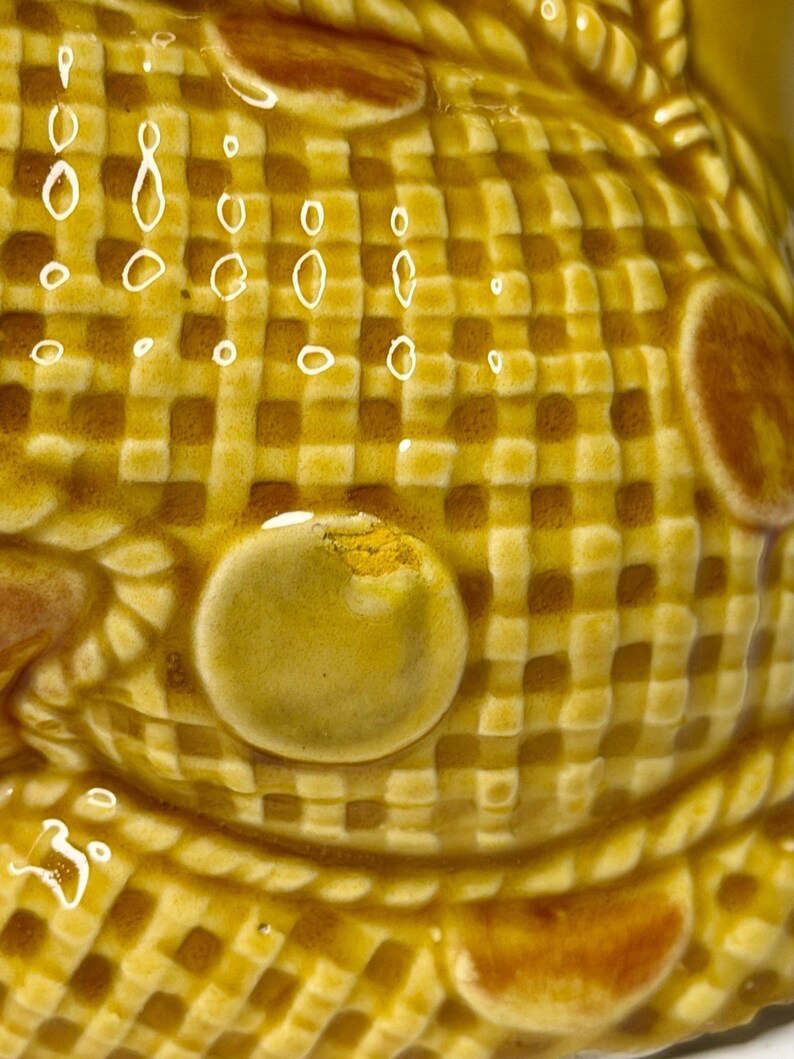 May include: Close-up of a ceramic object with a textured, checkered pattern in shades of yellow and brown. The surface has a glossy finish with raised, rounded details and a few small, white, droplet-shaped accents. The overall design suggests a decorative or functional item.