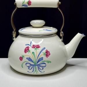May include: Cream-colored enamel teapot with a floral design. The teapot features a bouquet of pink flowers, blue ribbons, and green leaves. It has a white handle with a floral pattern and a metal handle. The spout and lid are trimmed with blue.