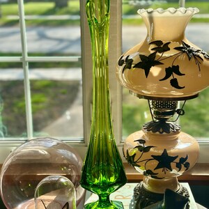 May include: A tall, green glass vase with a flared top and a geometric base. The vase is sitting on a table with a book, a glass dome with a butterfly inside, and a lamp with a white shade decorated with black floral designs.