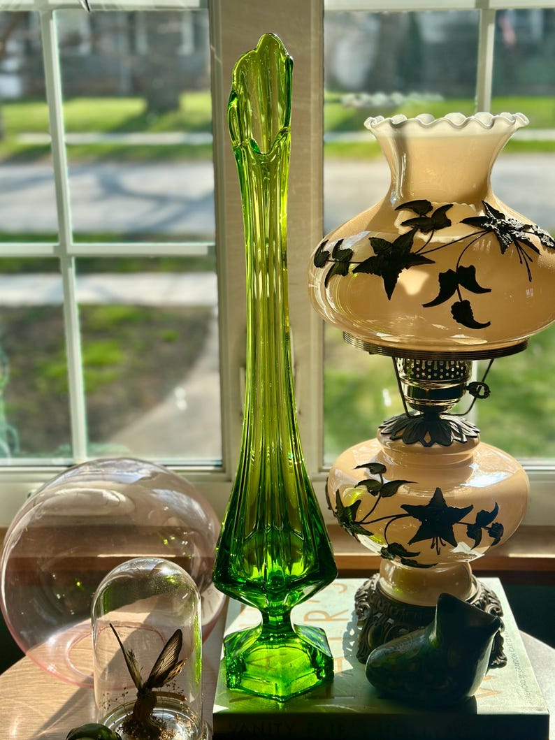 May include: A tall, green glass vase with a flared top and a geometric base. The vase is sitting on a table with a book, a glass dome with a butterfly inside, and a lamp with a white shade decorated with black floral designs.