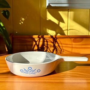 May include: White ceramic casserole dish with a blue floral design. The dish has a handle on one side.