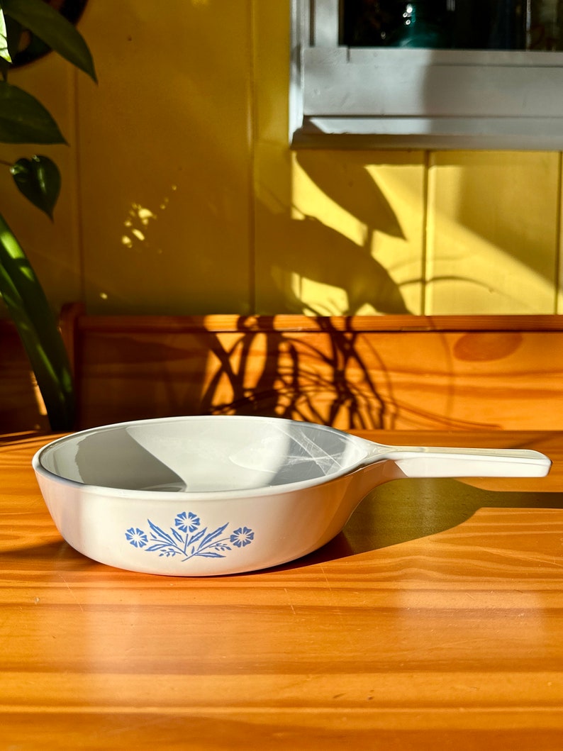 May include: White ceramic casserole dish with a blue floral design. The dish has a handle on one side.