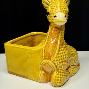 May include: A ceramic giraffe planter in shades of yellow and brown. The giraffe is in a seated position with a basket-like container attached. The giraffe has a checkered pattern and is a decorative item.