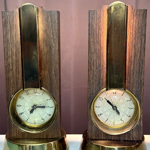 May include: Two vintage brass and wood clocks with a unique design. The clocks have a brass frame with a wood panel and a brass base. The clock faces are white with black numbers and hands.
