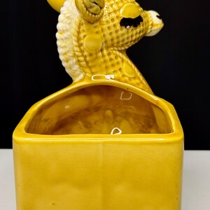 May include: A vintage ceramic planter in the shape of a giraffe's head, with a rectangular base. The giraffe is yellow with white accents, and the planter has a glossy finish. The planter is a decorative item for home decor.