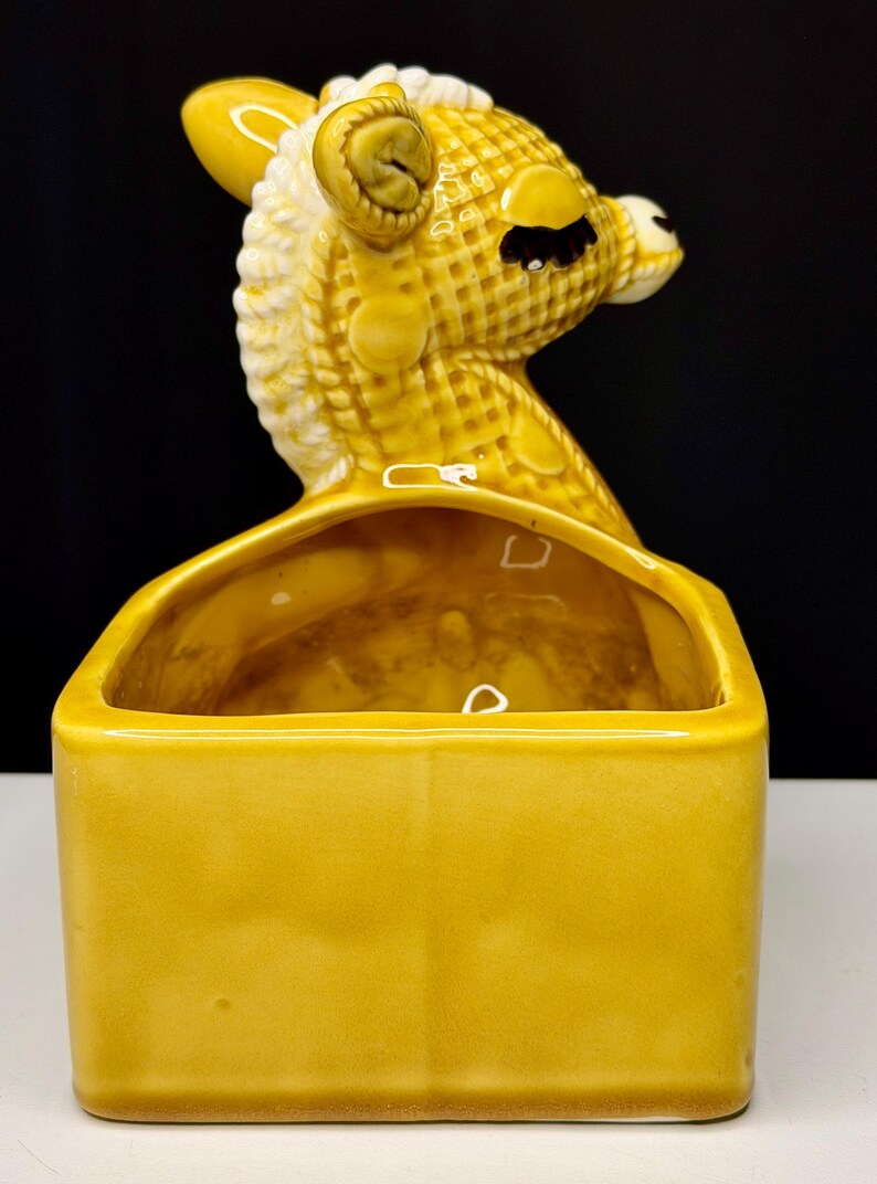 May include: A vintage ceramic planter in the shape of a giraffe's head, with a rectangular base. The giraffe is yellow with white accents, and the planter has a glossy finish. The planter is a decorative item for home decor.