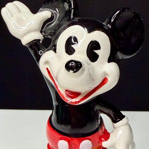 K&ouml;nnte beinhalten: Eine Keramikfigur von Mickey Mouse, einer klassischen Disney-Figur. Mickey wird mit einem schwarzen K&ouml;rper, wei&szlig;en Handschuhen und roten Shorts mit wei&szlig;en Kn&ouml;pfen dargestellt. Sein Gesicht ist wei&szlig; mit schwarzen Augen und einem roten L&auml;cheln. Er winkt mit einer Hand.