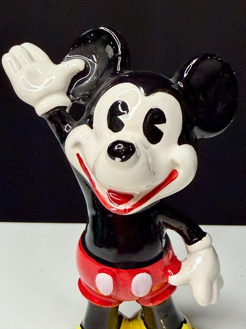 K&ouml;nnte beinhalten: Eine Keramikfigur von Mickey Mouse, einer klassischen Disney-Figur. Mickey wird mit einem schwarzen K&ouml;rper, wei&szlig;en Handschuhen und roten Shorts mit wei&szlig;en Kn&ouml;pfen dargestellt. Sein Gesicht ist wei&szlig; mit schwarzen Augen und einem roten L&auml;cheln. Er winkt mit einer Hand.