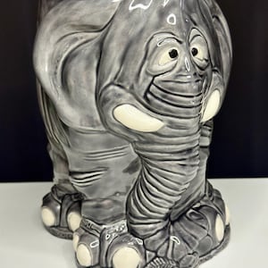 May include: Gray ceramic elephant-shaped planter with a smiling face and large ears. The planter has a detailed trunk, tusks, and feet. The elephant is painted in shades of gray with white accents.