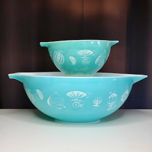 May include: Two vintage turquoise mixing bowls stacked on top of each other. The bowls have a white design of hot air balloons and other whimsical elements. The larger bowl is a brighter turquoise than the smaller bowl.