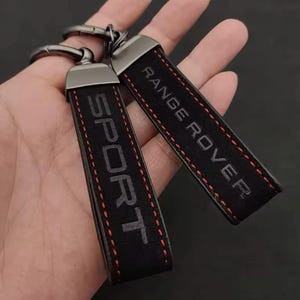 Könnte beinhalten: Zwei schwarze Leder-Schlüsselanhänger mit den geprägten Wörtern "SPORT" und "RANGE ROVER" in Grau. Die Schlüsselanhänger haben rote Nähte und silberfarbene Metallbeschläge.