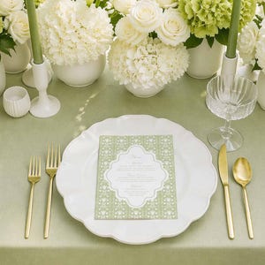 May include: A beautifully set table with white flowers, green candles, and gold cutlery. A white plate with a decorative menu card sits in the center. The table is covered with a light green tablecloth, creating an elegant and inviting atmosphere.
