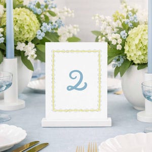 Könnte beinhalten: Eine Tischdekoration mit hellblauer Tischdecke, weißen Tellern und goldenem Besteck. Eine weiße Tischkarten mit der blauen Zahl 2 steht im Mittelpunkt. Blaue Kerzen und Blumengestecke ergänzen die elegante Einrichtung.