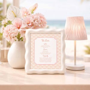 May include: A white framed menu with a pink and white design, listing wine, beer, and spirits. A white vase with pink flowers and a small white lamp are on a table. The background shows a beach scene.
