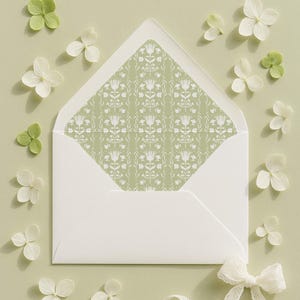 Op de afbeelding: Een witte envelop met een bloemmotief aan de binnenkant, omringd door witte en groene bloemblaadjes en een wit kanten lint. De binnenkant van de envelop heeft een herhalend patroon van gestileerde bloemen en bladeren op een lichtgroene achtergrond.
