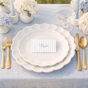 May include: Elegant table setting with white scalloped plates, gold cutlery, and a striped place card that reads "Baylor." The table is set on a light blue tablecloth, with floral arrangements and candles.
