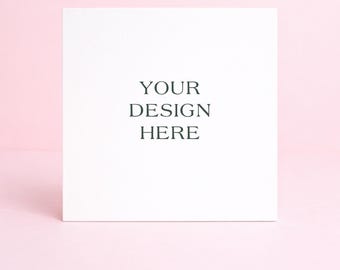 May include: A white square card with the text "YOUR DESIGN HERE" in a dark green font. The card is centered on a light pink background, creating a clean and minimalist aesthetic. Ideal for showcasing custom designs.