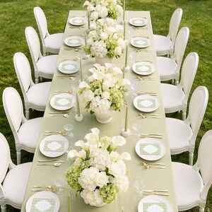 a beautifully set outdoor dining table with white chairs, white plates, and white flowers arranged in vases. The table is set for a formal event or celebration, with elegant tableware and floral centerpieces.