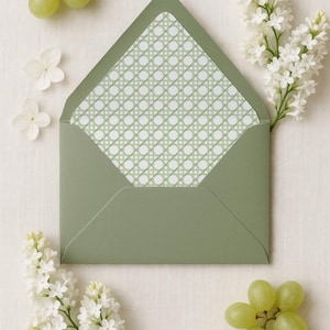 May include: An open sage green envelope featuring a white and light green cane webbing pattern on the interior liner, accompanied by clusters of green grapes and sprigs of white lilac flowers arranged around the stationery item.